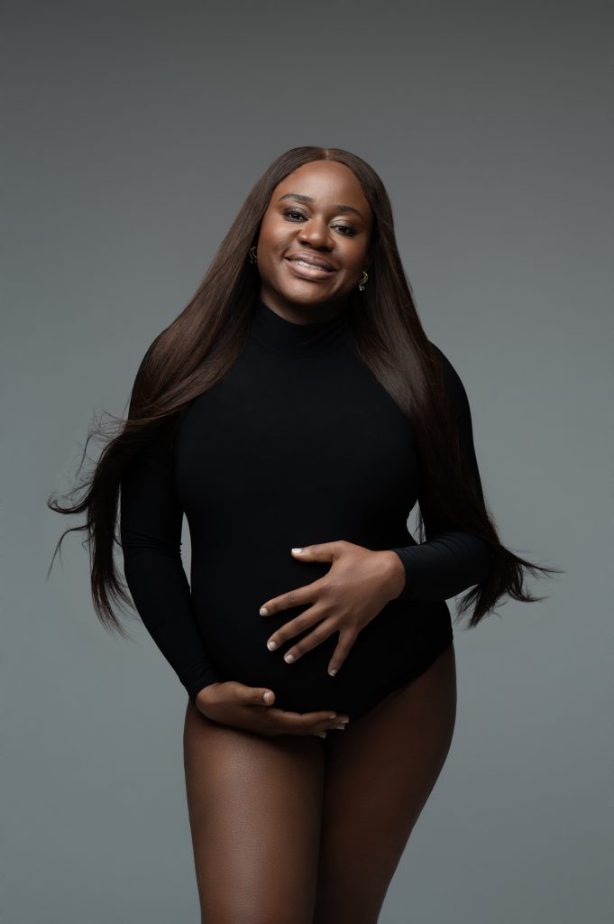 Pregnant woman, smiling and lovingly embracing her pregnancy belly. She has a black bodysuit on and long brown hair